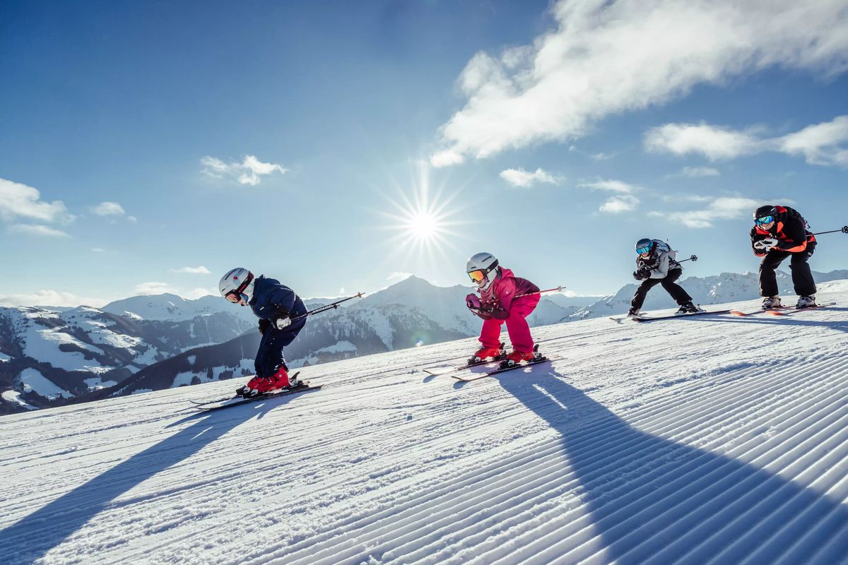 Vier Skifahrer, darunter zwei Kinder im Vordergrund, fahren eine präparierte Skipiste hinunter. Die Sonne strahlt hell am Himmel über verschneiten Bergen.