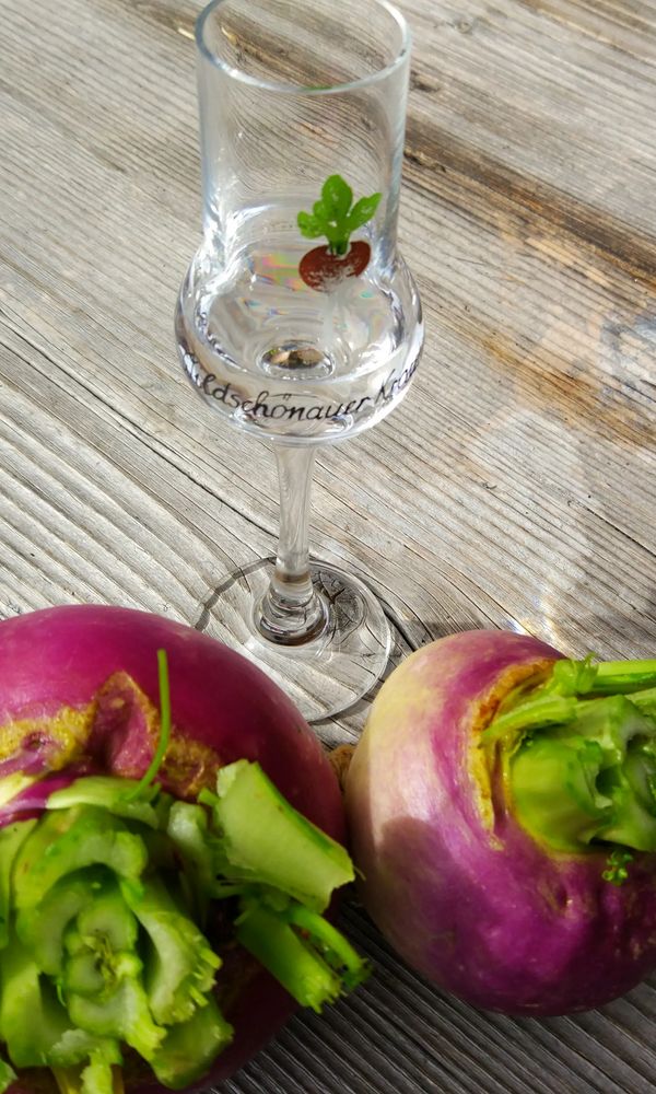 Zwei pinkfarbene Krautingerrüben liegen auf einem Holztisch neben einem Schnapsglas mit einem grünen Blatt.