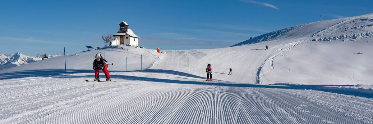 Eine verschneite Skipiste mit mehreren Skifahrern und einem kleinen, weißen Gebäude auf einem Hügel unter einem blauen Himmel.