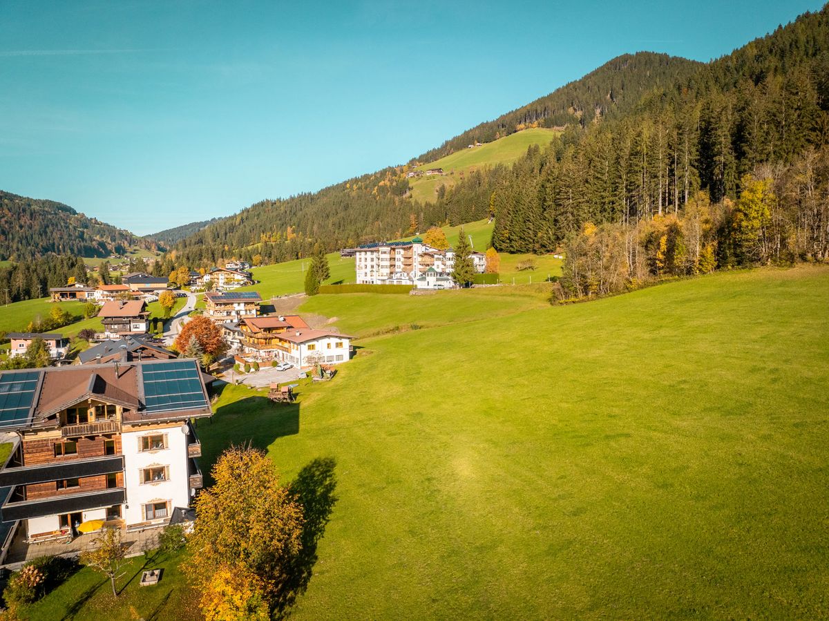 Eine Luftaufnahme eines Dorfes in einem Tal, umgeben von grünen Wiesen und bewaldeten Bergen. Einige Bäume zeigen Herbstfarben.