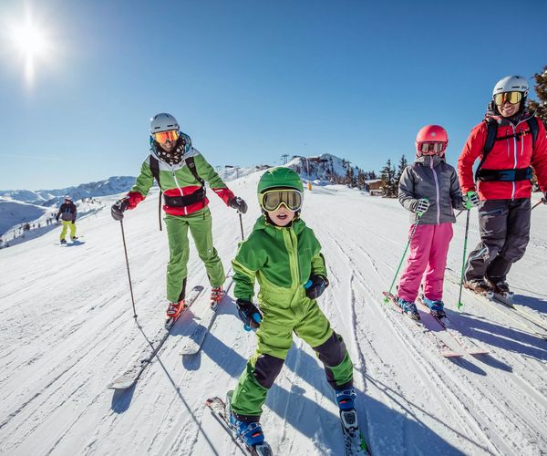 Eine Gruppe von vier Skifahrern, darunter Kinder und Erwachsene, steht auf einer präparierten Skipiste unter strahlendem Sonnenschein.