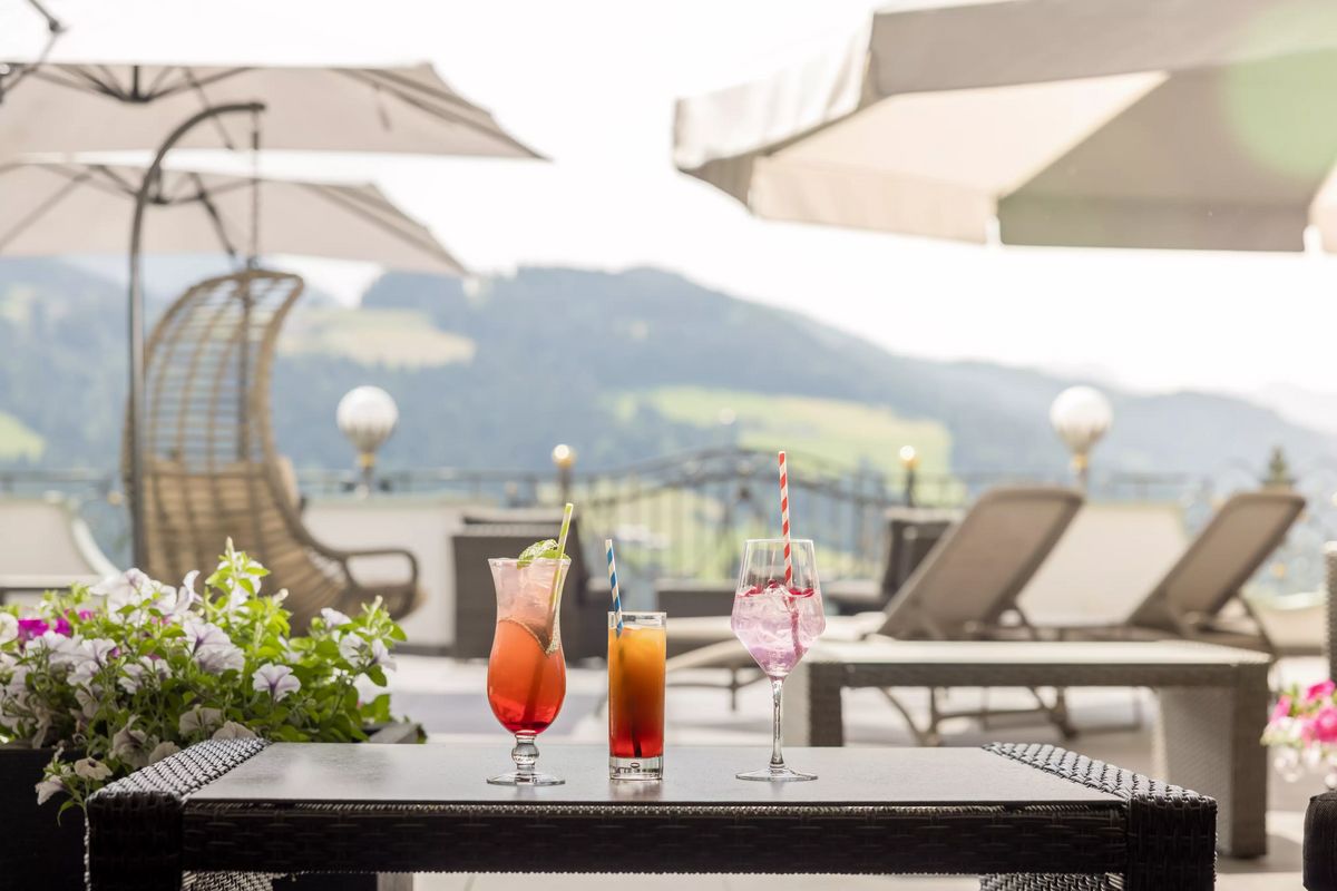 Drei bunte Cocktails stehen auf einem Tisch auf einer Terrasse mit Blick auf eine bergige Landschaft. Im Hintergrund sind Sonnenschirme und Liegestühle zu sehen.