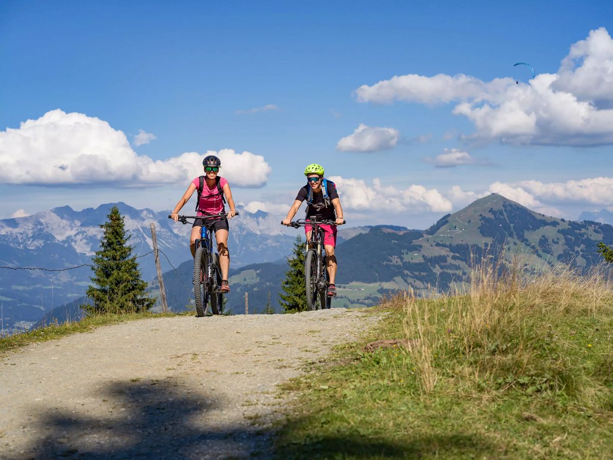 Zwei Personen fahren mit Mountainbikes einen Schotterweg bergauf. Im Hintergrund sind bewaldete Berge und ein blauer Himmel mit Wolken zu sehen, sowie ein Paraglider.